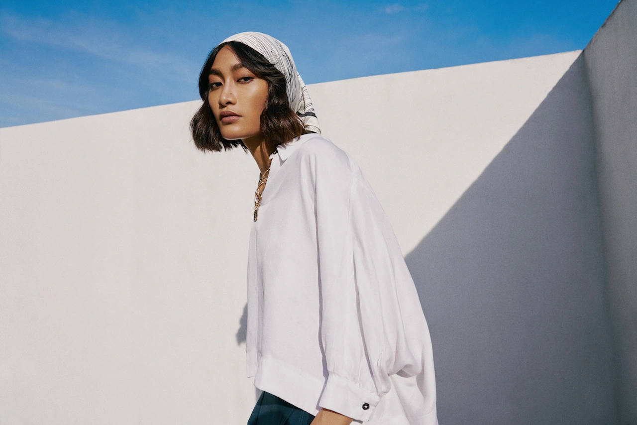 Fashion editorial shot of a model in a headscarf and white blouse against a sunlit wall